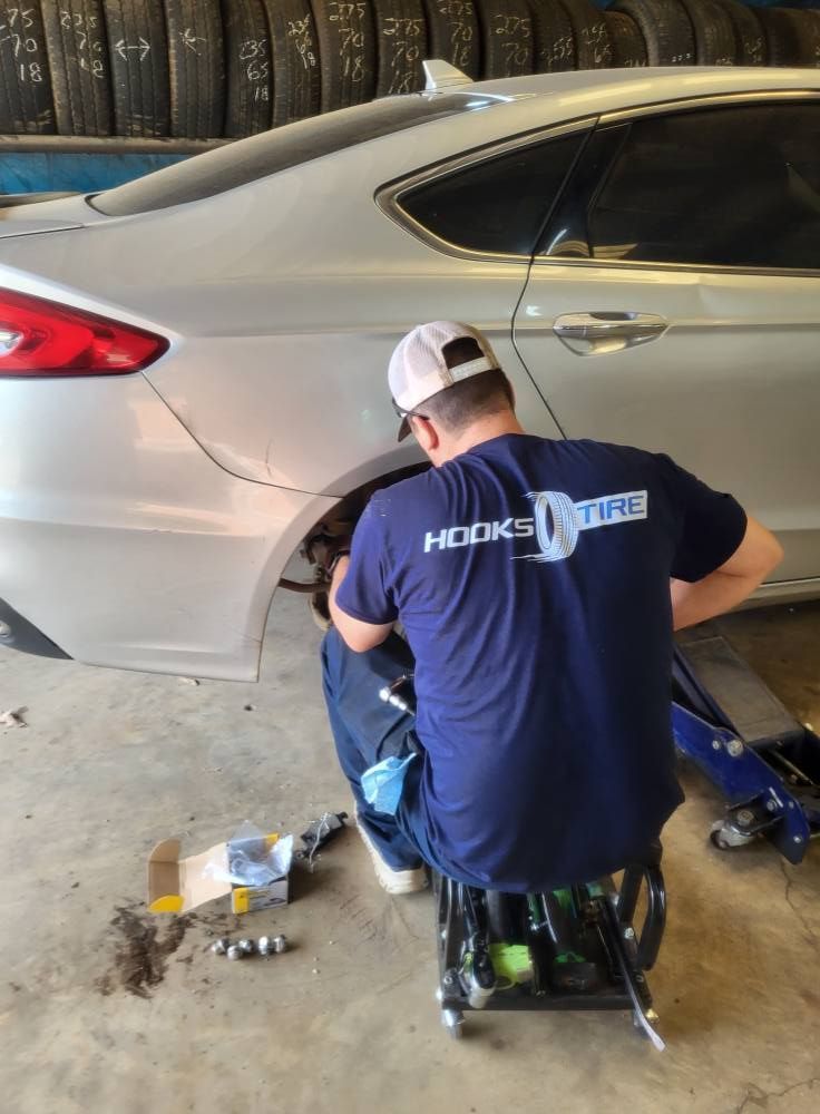 Mechanic working under a silver sedan in a garage, surrounded by tools and car parts.