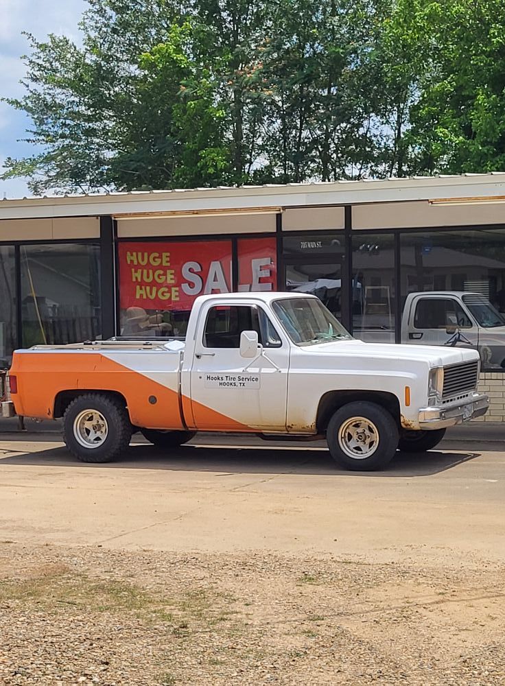 White and orange vintage pickup truck parked outside a storefront with a red sale sign