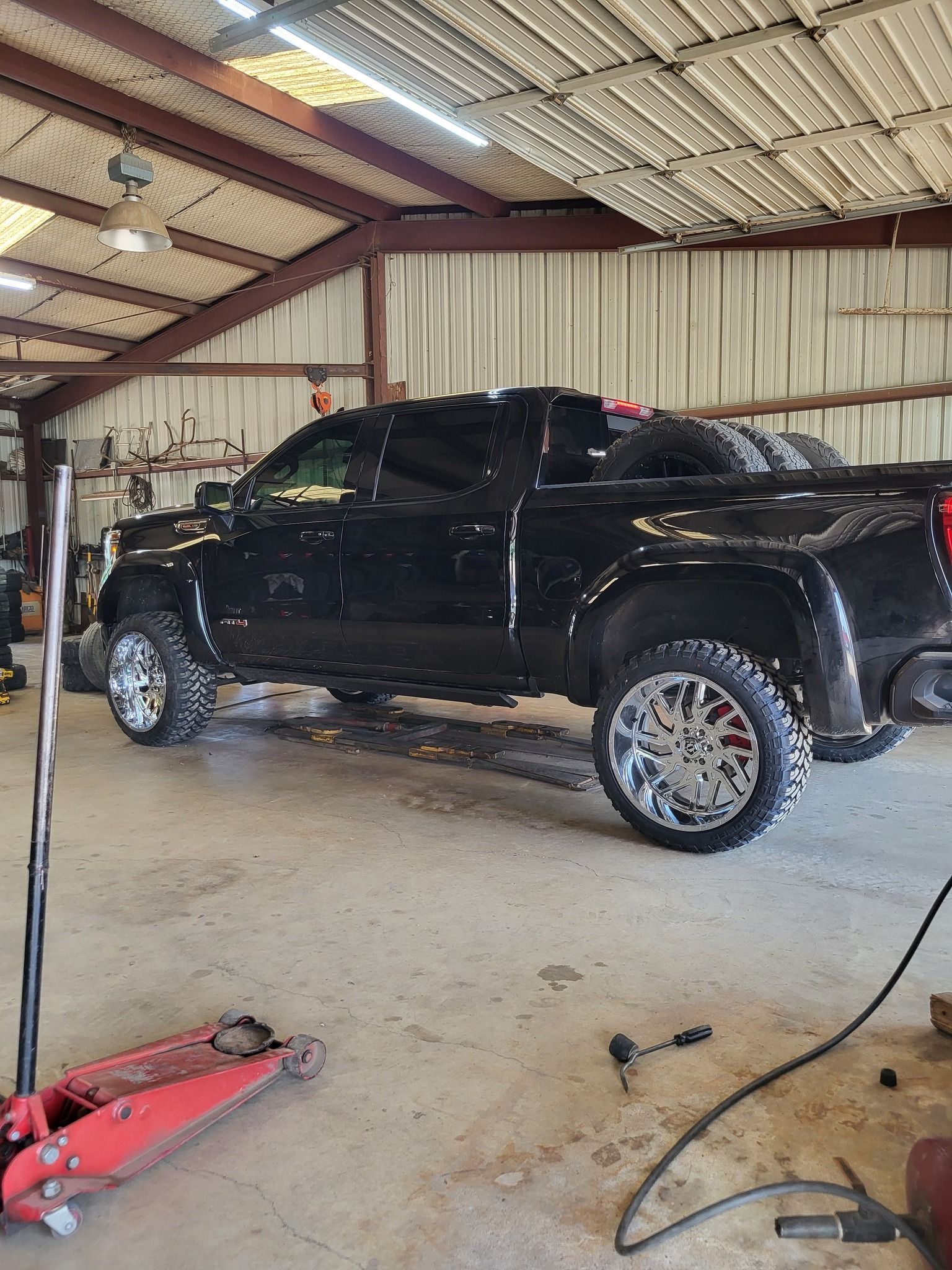 Black pickup truck in a garage with a red floor jack and tools on the concrete floor.