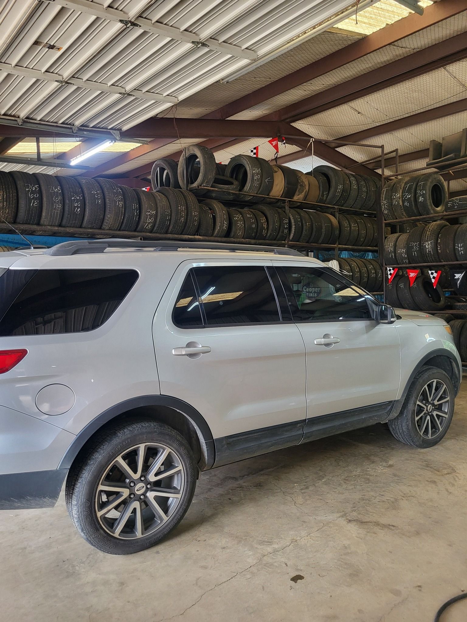 Silver SUV parked in an auto repair garage under stacked tires on a mezzanine shelf.