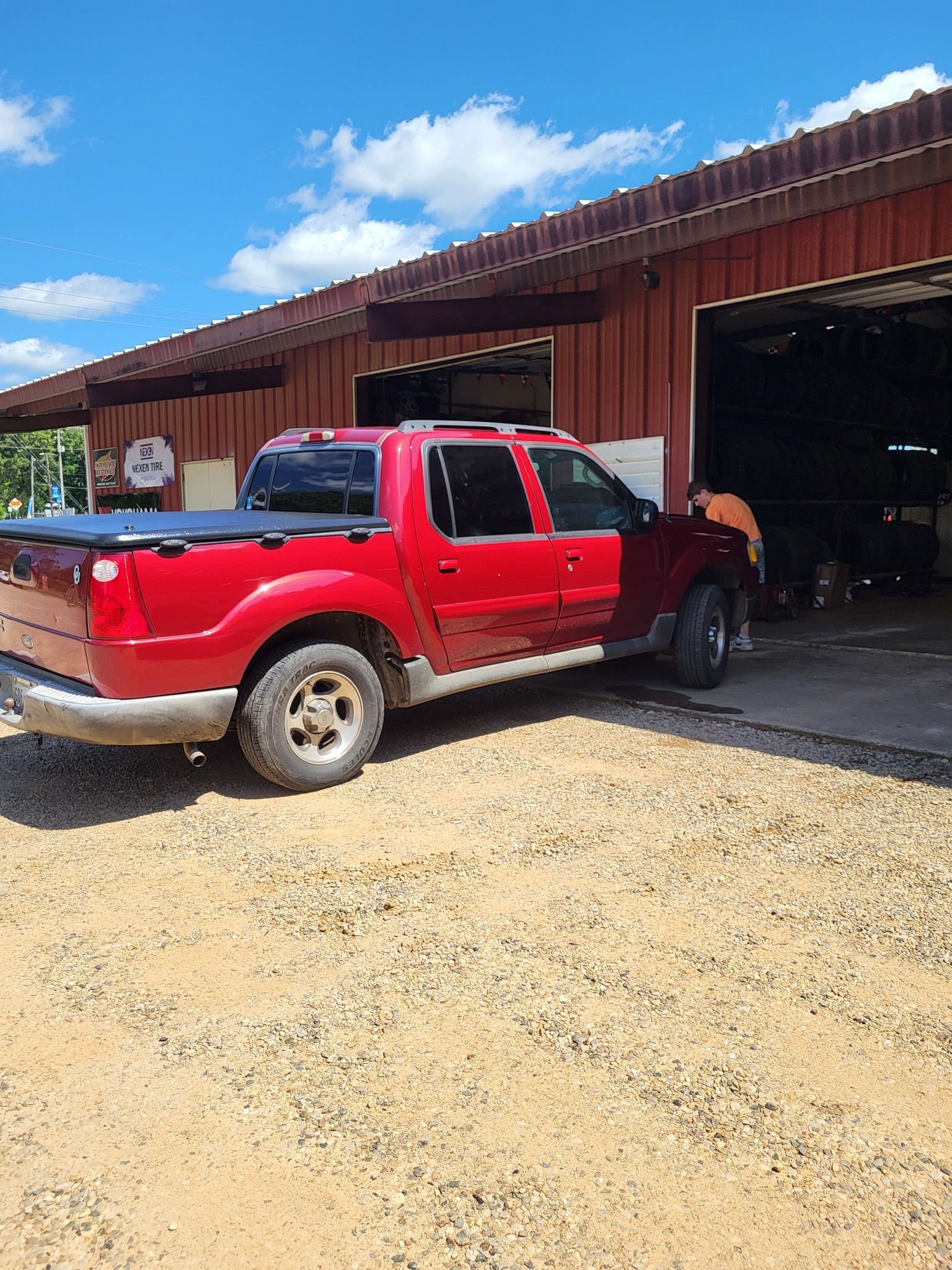 Red pickup truck parked beside a barn with an open garage door on a sunny gravel lot