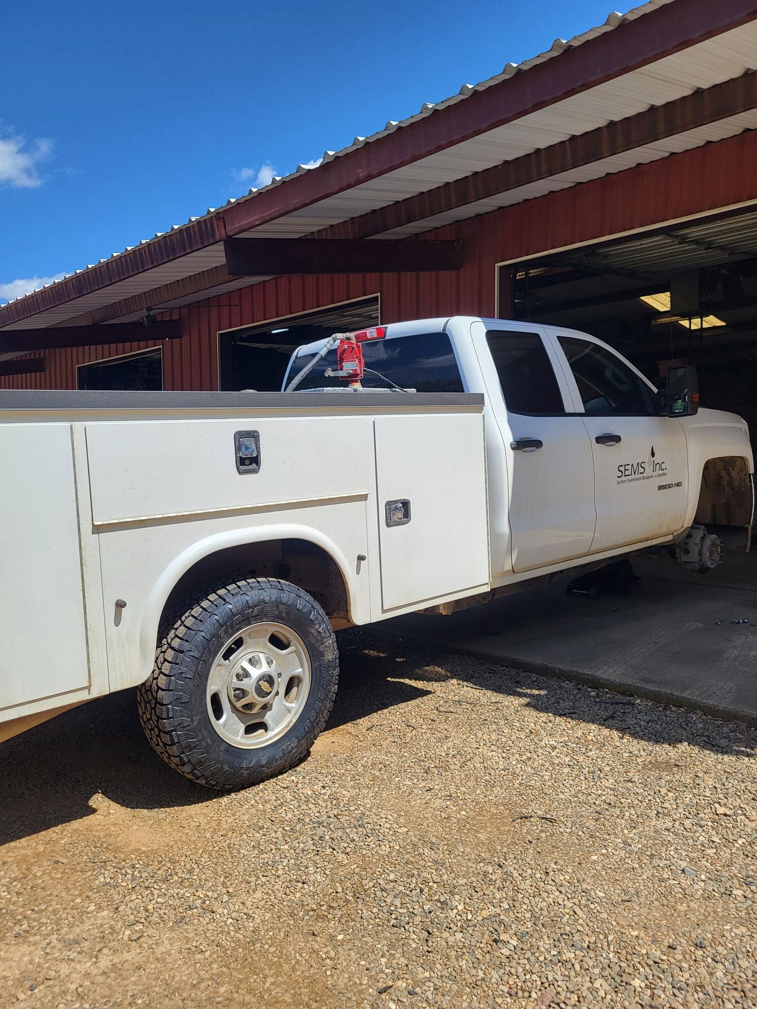 White utility truck parked under a barn roof on a gravel driveway.