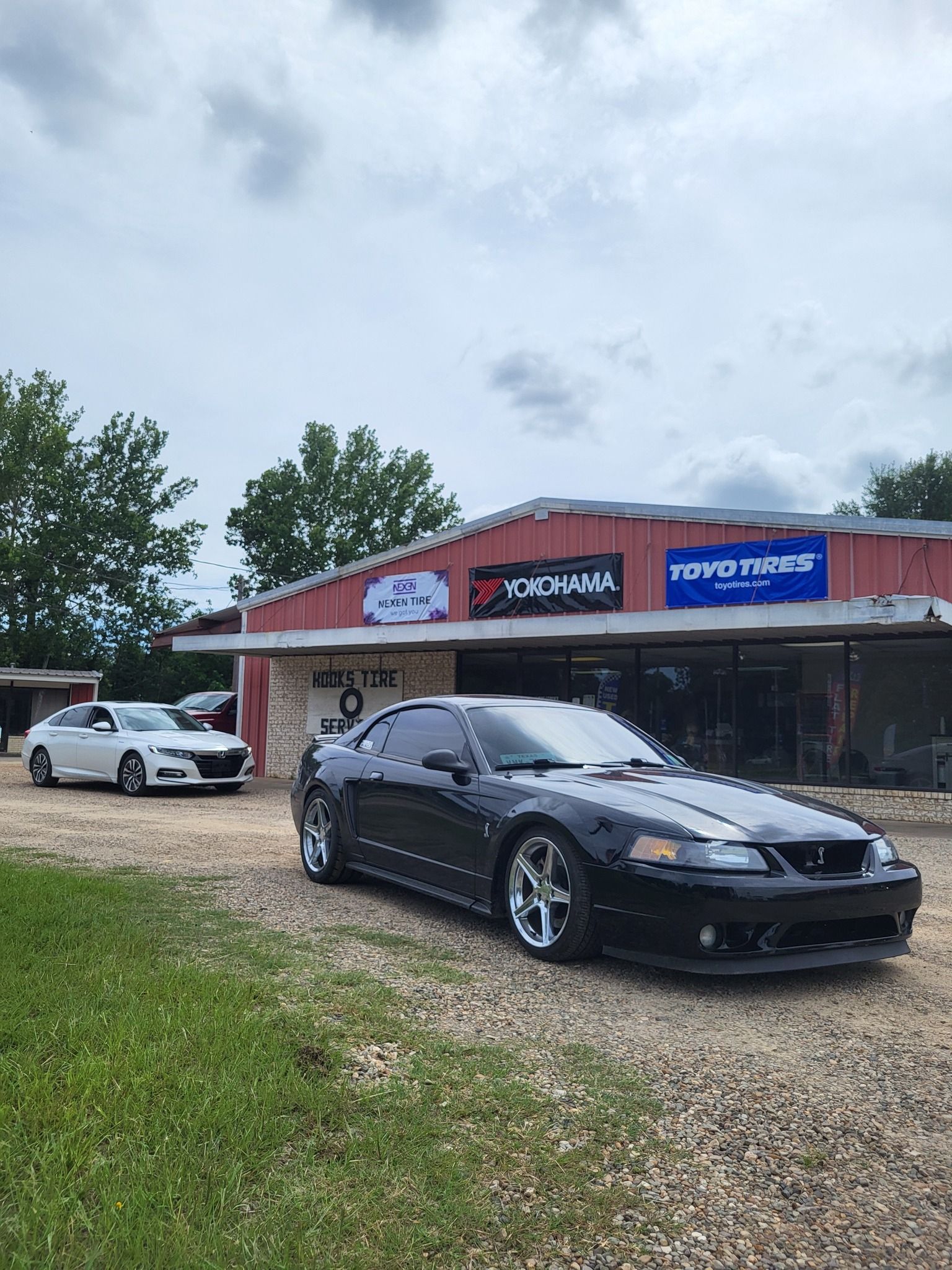 Black sports car parked outside a small store with a white SUV and cloudy sky overhead