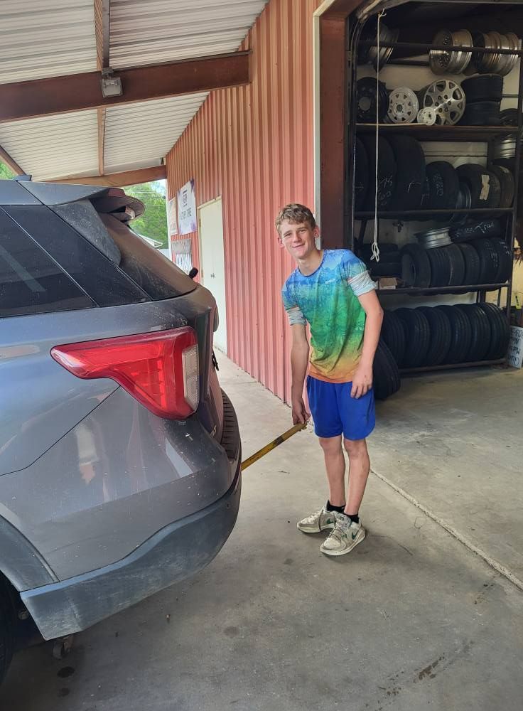 Smiling child standing beside a gray SUV at a garage, holding a hose near stacked tires.