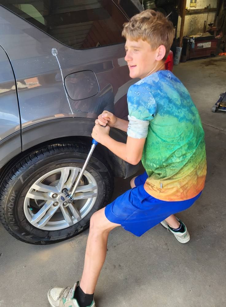Boy using a pump on a car tire in a garage, wearing a blue-and-green shirt and shorts.