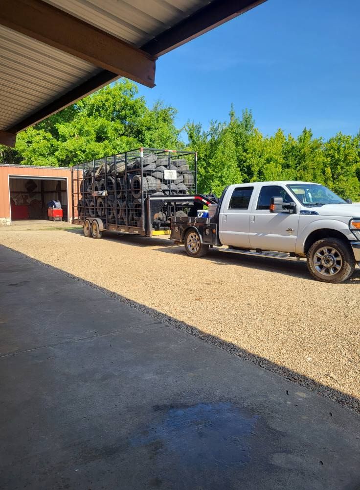 Silver pickup truck towing a black trailer in a gravel driveway beside a red building and green trees