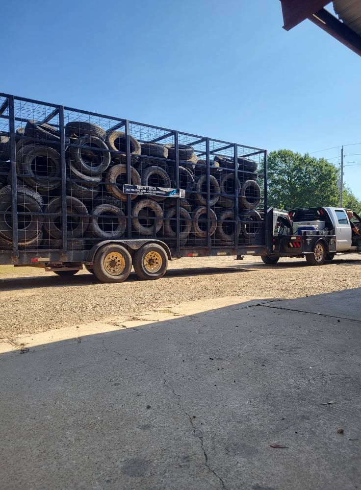 Truck hauling a trailer stacked with tires on a sunny street, with a tow vehicle attached.