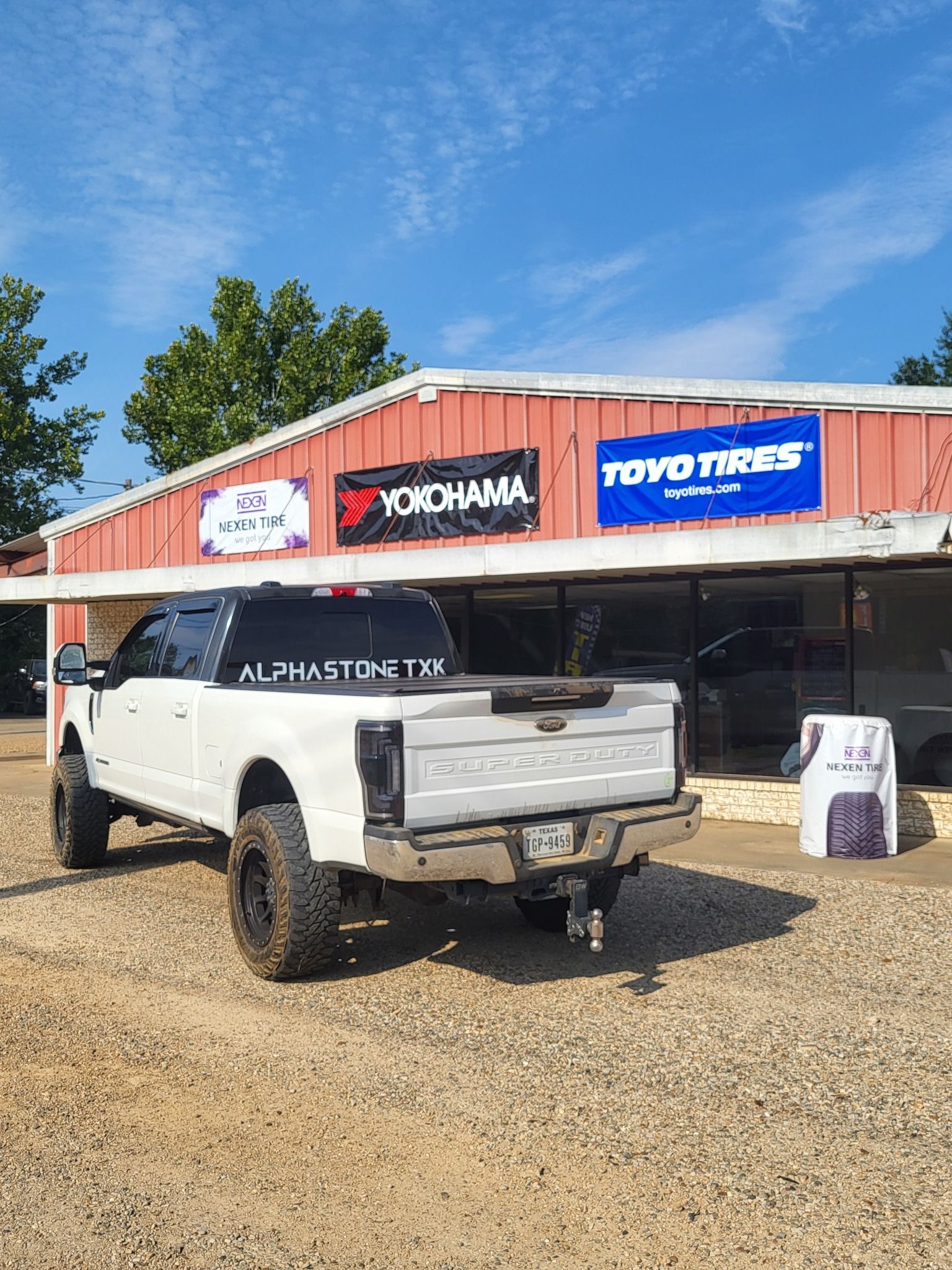 White pickup truck parked outside a red tire shop with YOKOHAMA and Toyo Tires signs.