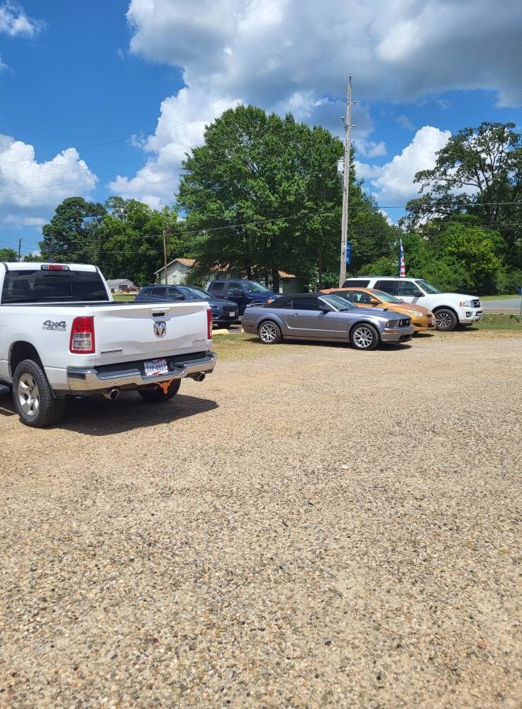 Parking lot with several cars and pickup trucks under a blue sky with clouds and trees nearby