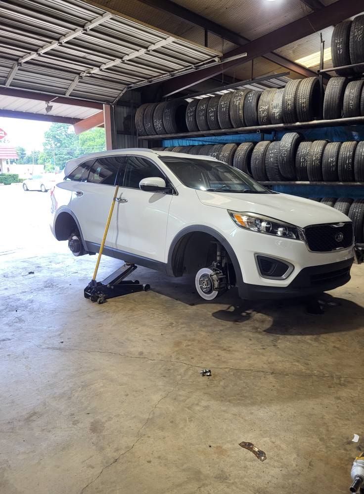 White SUV raised on jack in auto shop with tires stacked along the wall and a tire tool nearby