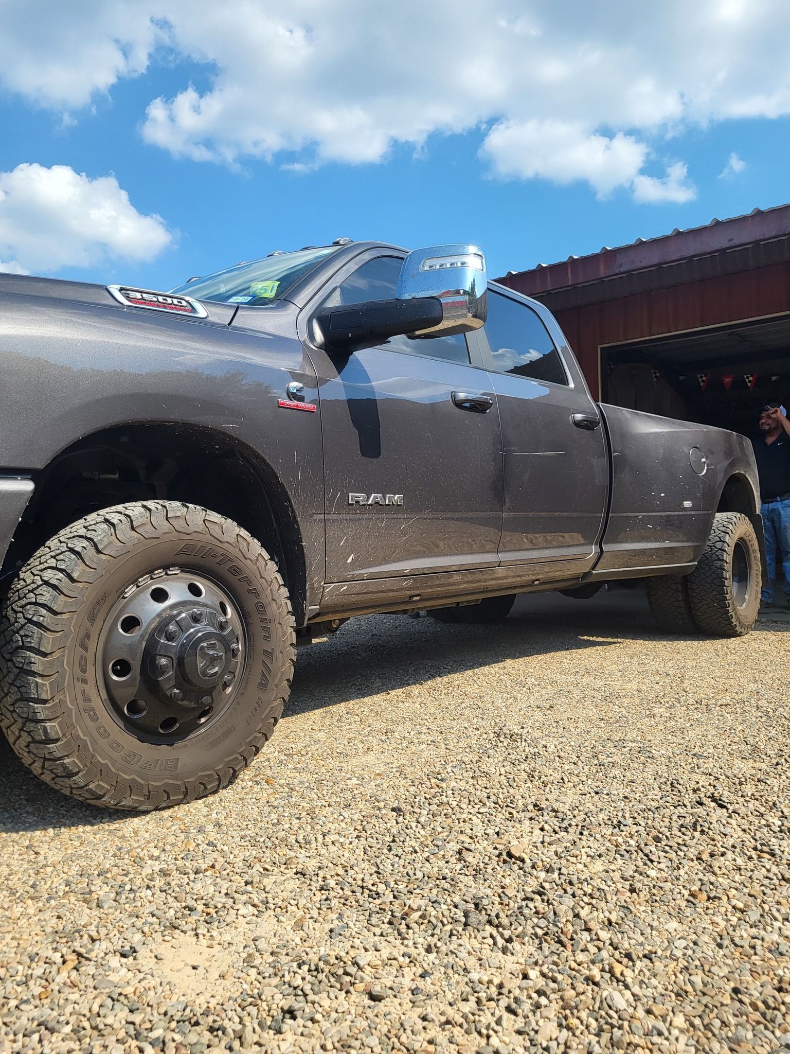 Mud-covered gray pickup truck parked on gravel beside a red-roofed building under a blue sky.