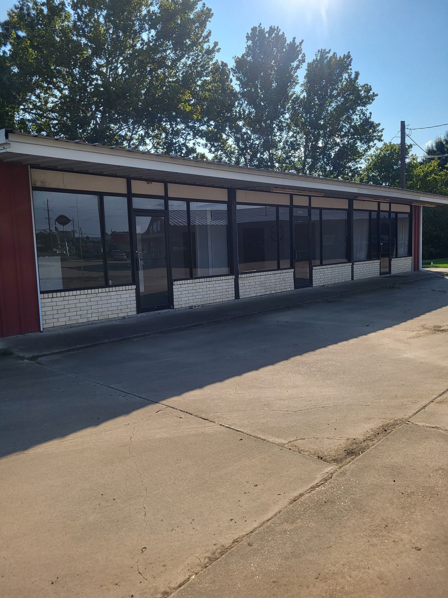 Single-story storefront with dark awning and glass windows beside a sunlit road, trees behind it.