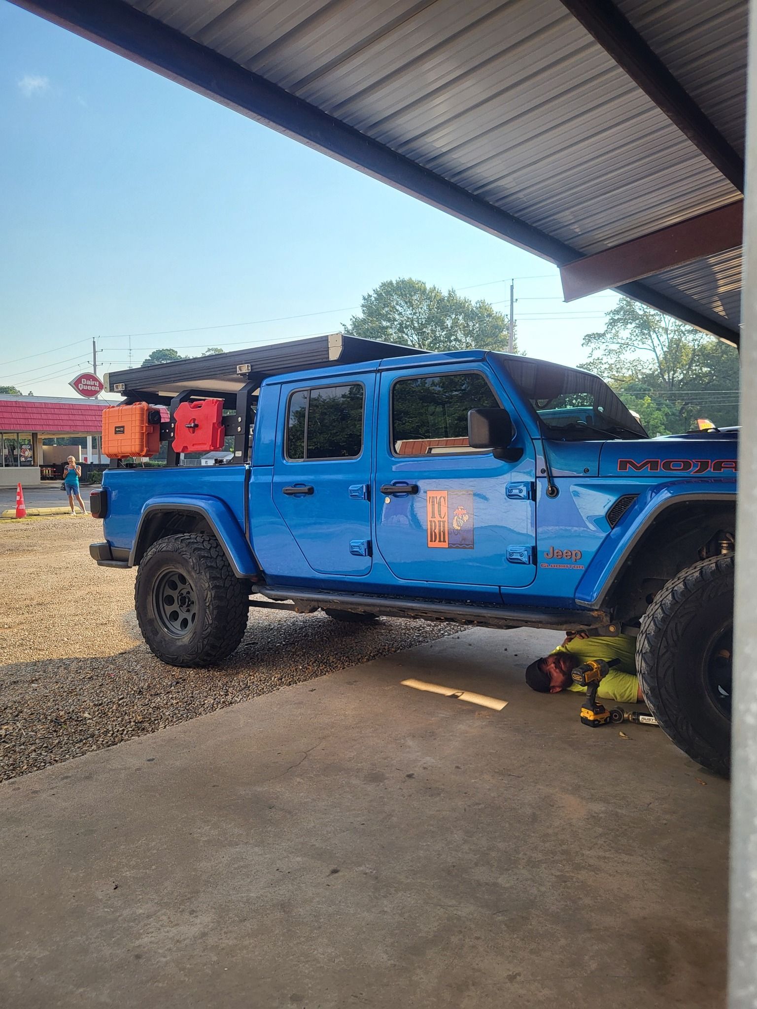 Blue off-road pickup truck parked under a roofed area, with a spare tire and gear in the back.
