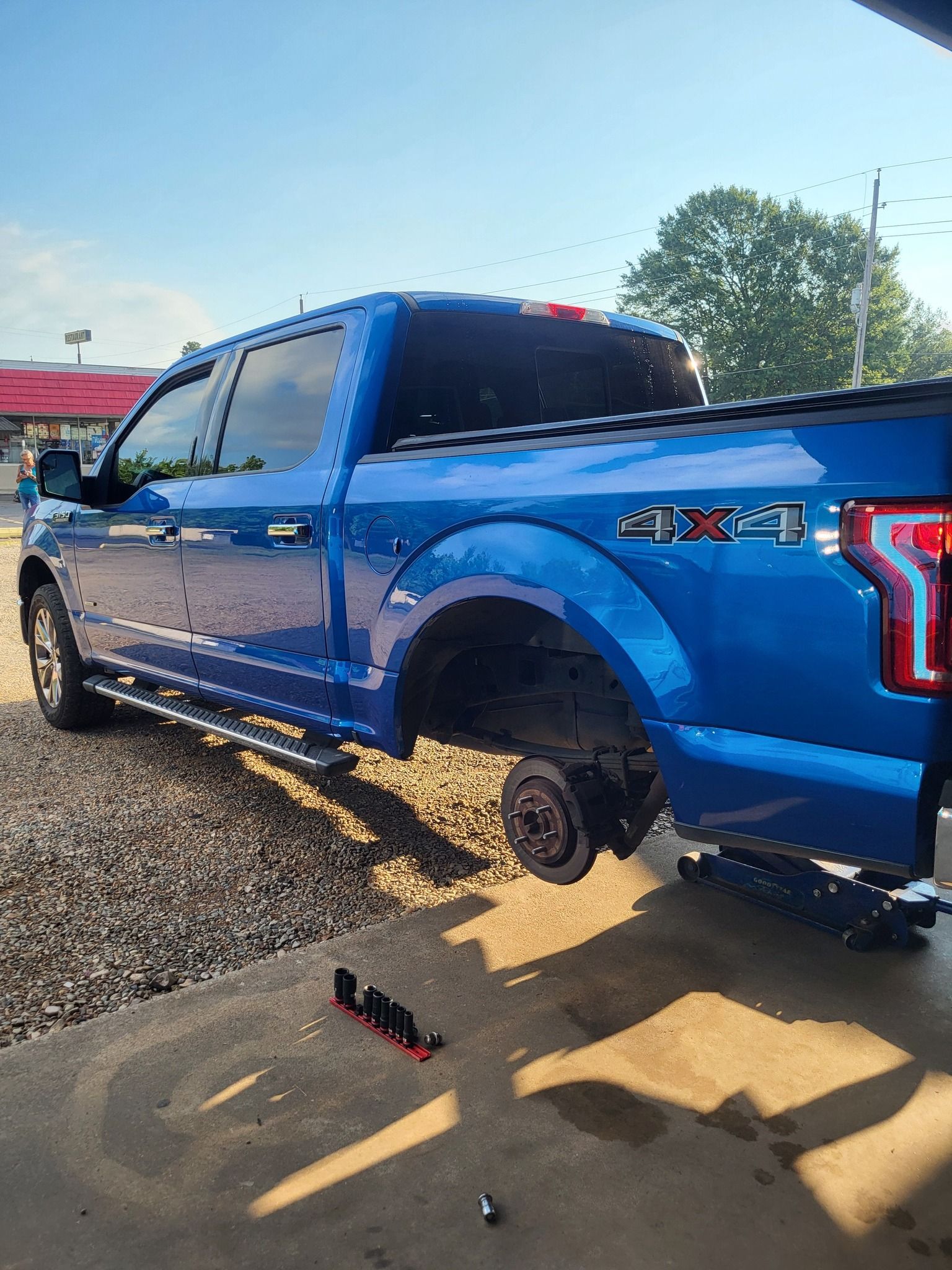 Blue pickup truck with rear wheel removed, parked on gravel in bright sunlight.