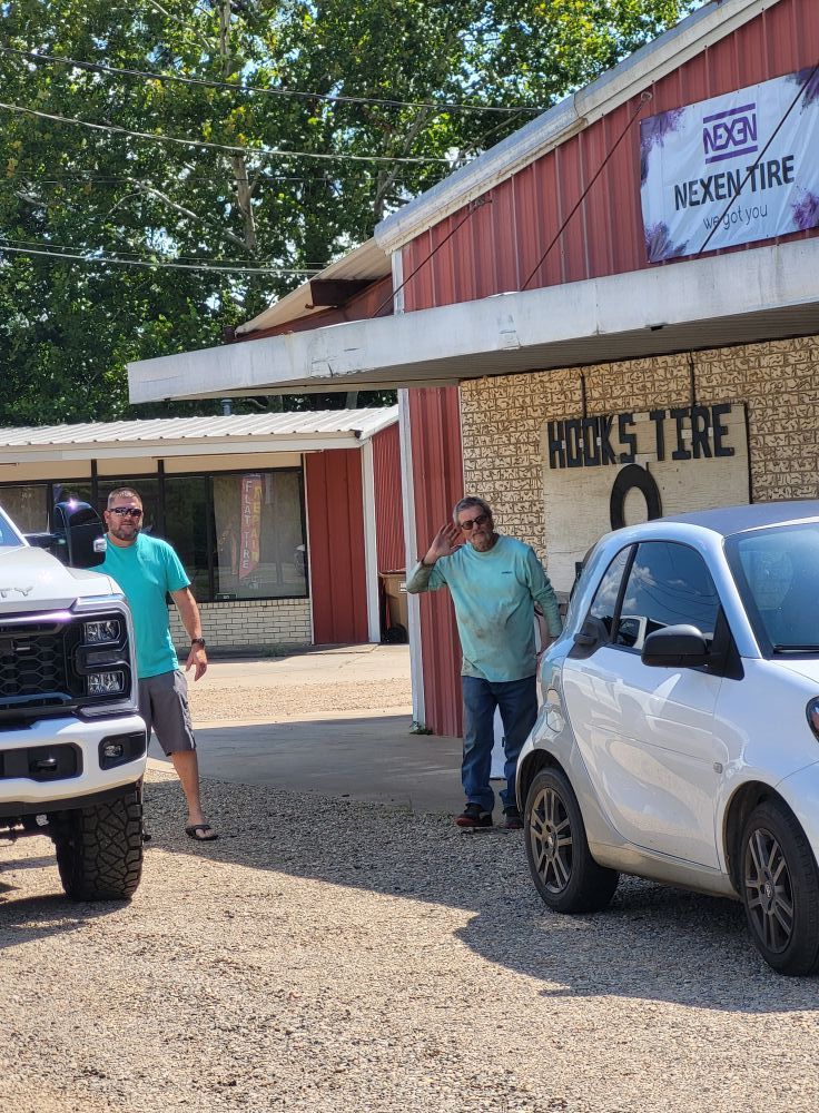 People outside a red building with a tire shop sign, beside two parked trucks and a white SUV.