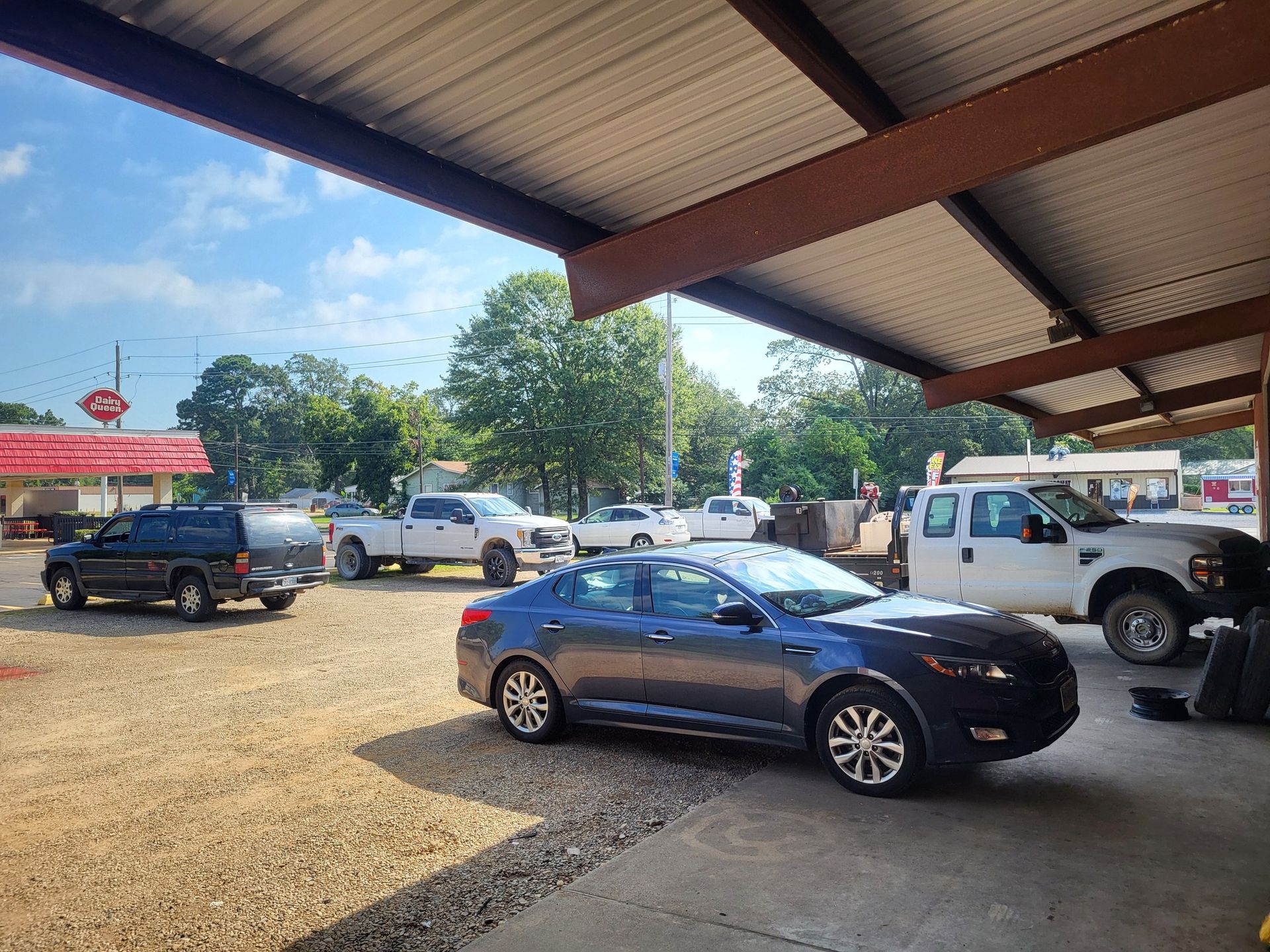 Cars parked under a metal-roofed carport on a sunny day, including a dark sedan in the foreground.