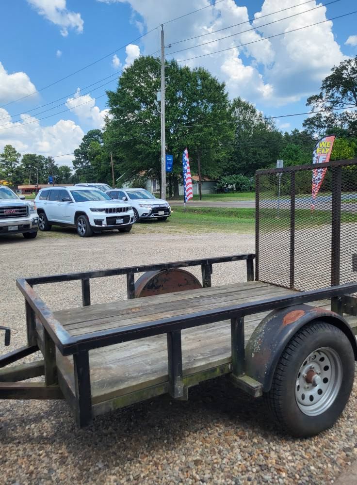 Utility trailer parked beside a chain-link fence on a gravel lot, with vehicles and trees in the background