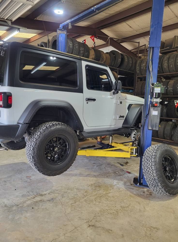 White Jeep Wrangler on a lift in an auto repair shop, with a detached wheel in the foreground.