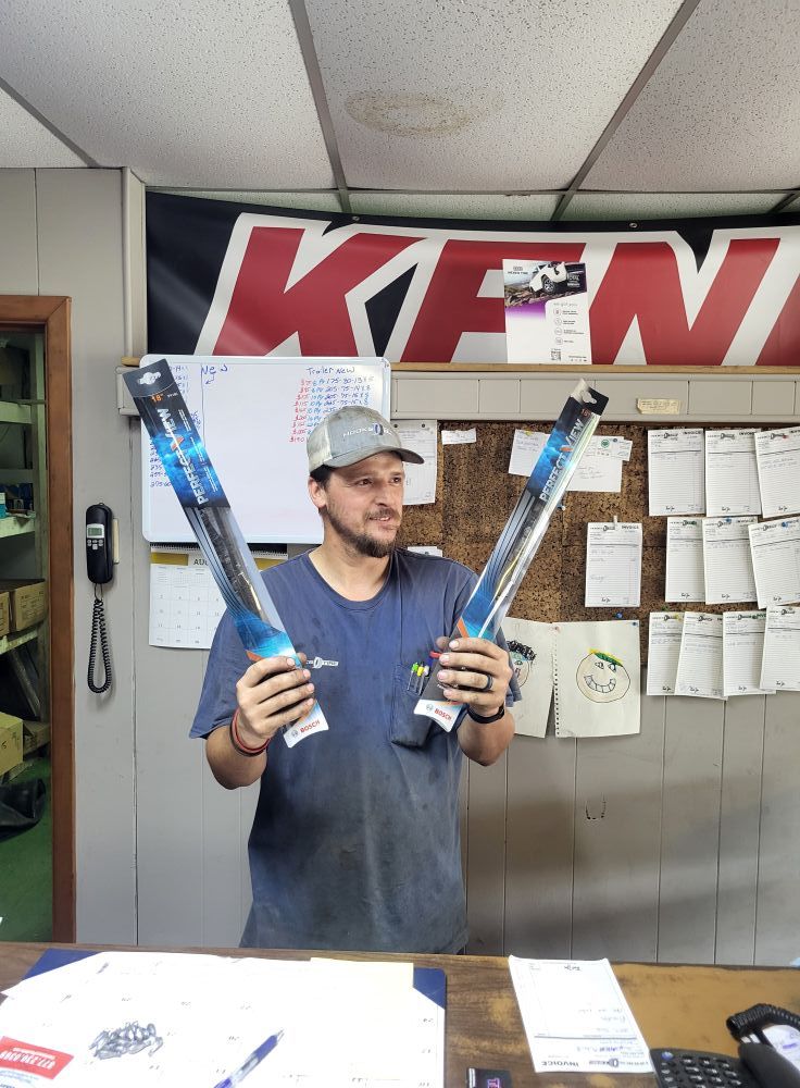 Man holding two long blue tools in a workshop, with papers and a large sign on the wall.