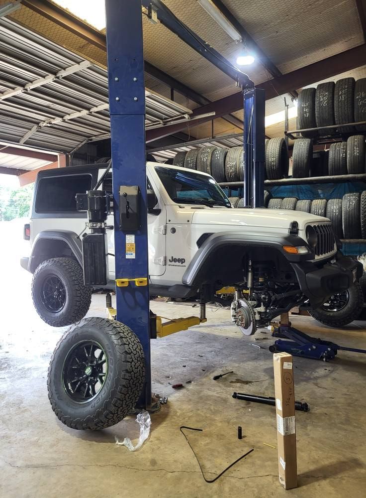 White Jeep lifted in a garage on a hydraulic hoist, with tires stacked along the wall and a detached wheel in front.