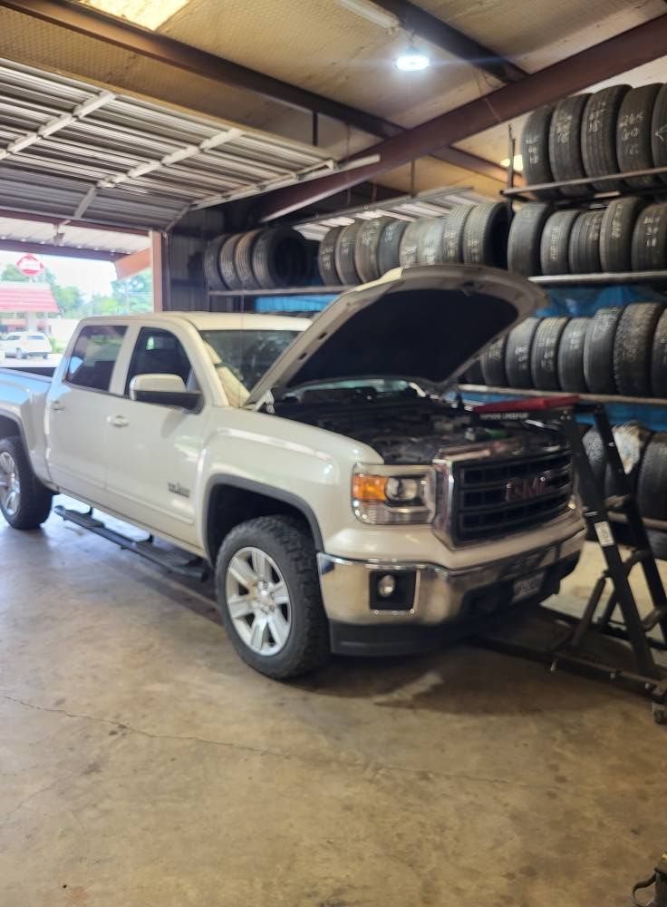 Silver pickup truck with hood open inside a tire shop, tires stacked on wall shelves.