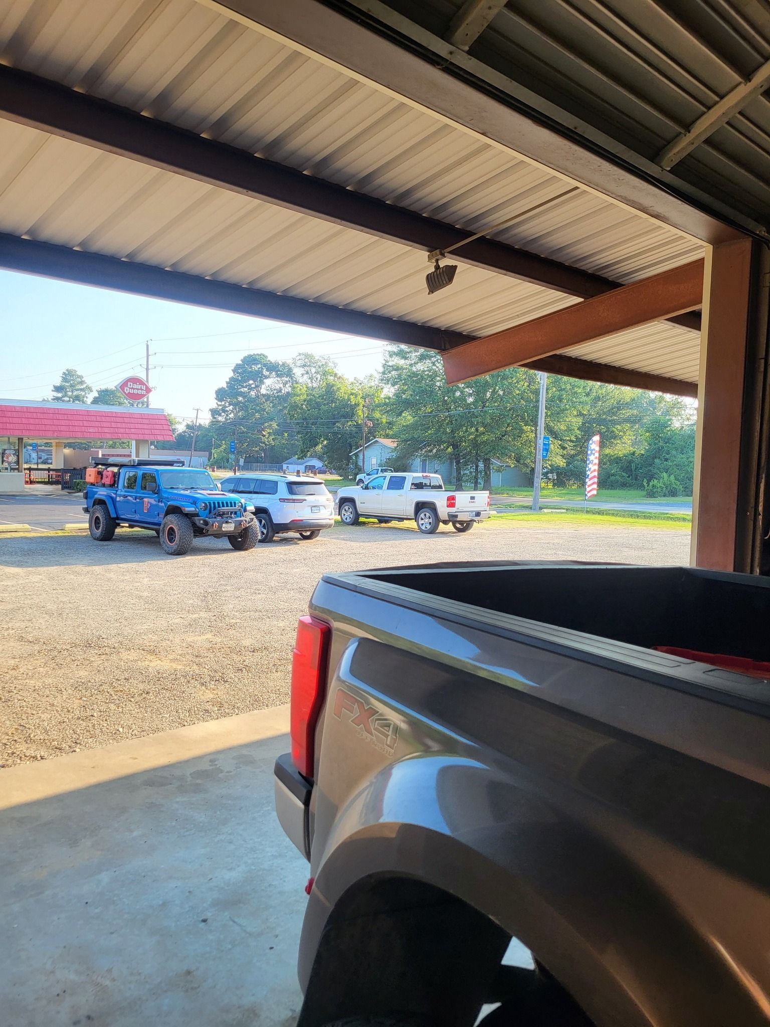 Parking lot view from a shaded carport, with blue and white pickup trucks near a storefront.