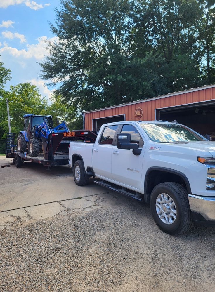 White pickup truck towing a blue tractor on a trailer beside a red barn on a sunny day