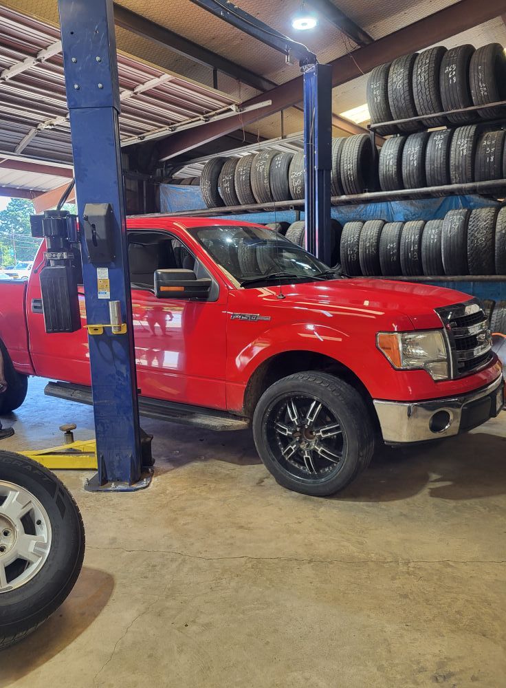 Red pickup truck on a lift in an auto repair shop, with stacks of tires in the background.