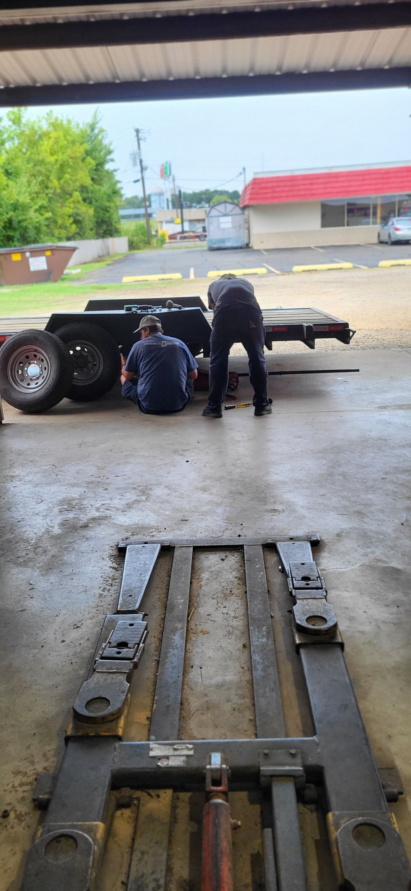 Two workers crouch by a black vehicle in a wet garage bay, with a metal ramp in the foreground.
