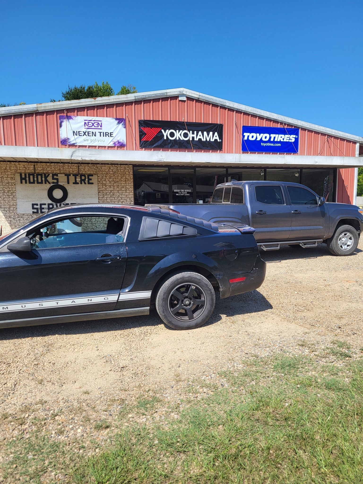 Black car and dark pickup parked outside a shop with Yokohama and Powertrac signs under a blue sky
