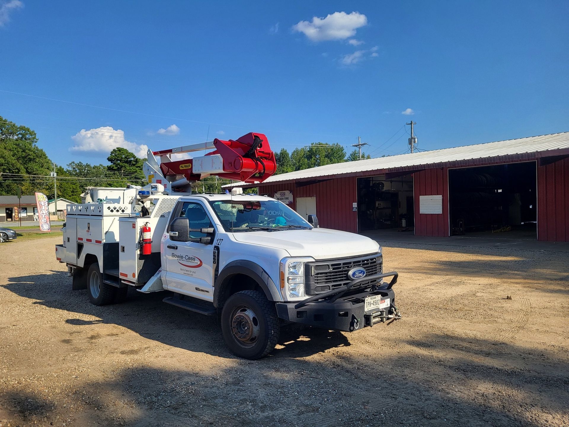 White utility truck with red crane parked on a dirt lot beside open red garage bays under a blue sky.