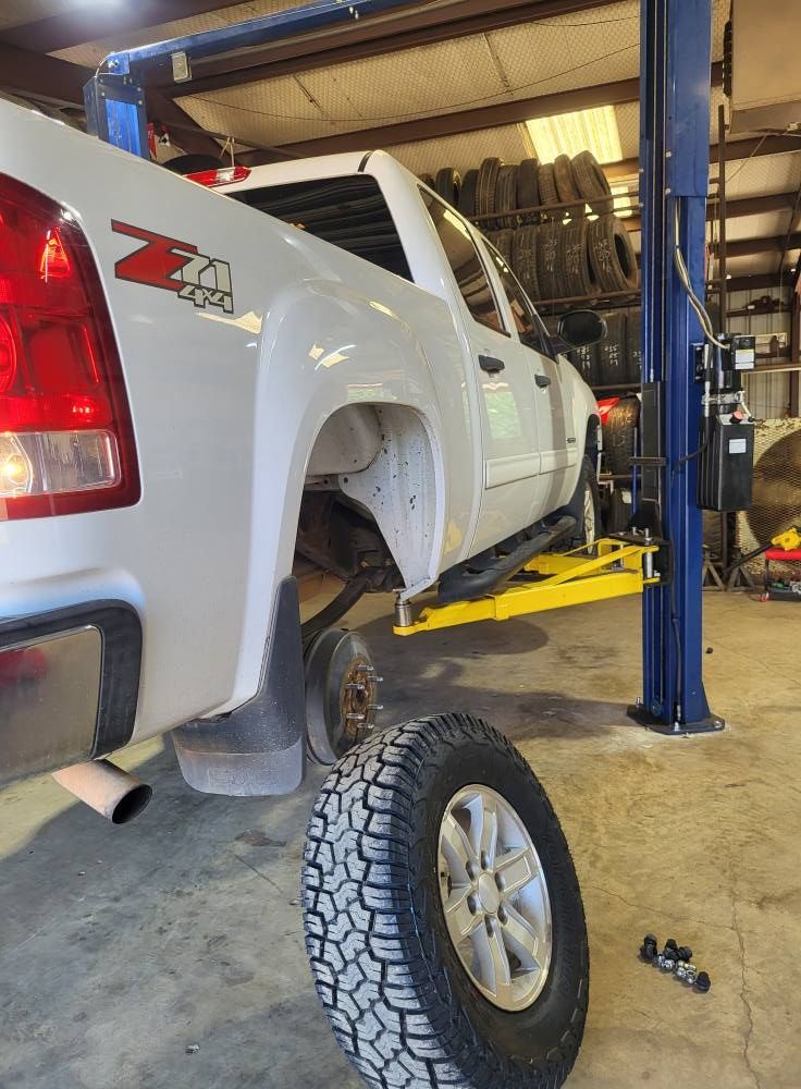 White pickup truck lifted in an auto shop with a detached wheel on the floor and a mechanic visible underneath