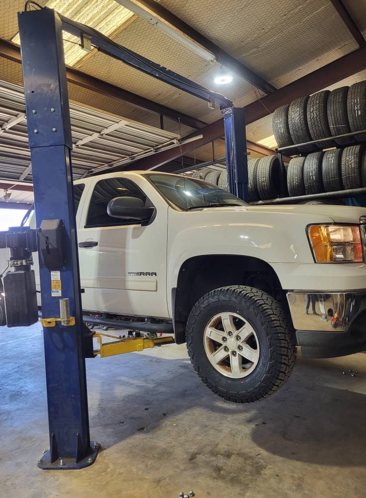 White pickup truck lifted on a blue car lift inside an auto repair shop with stacked tires.