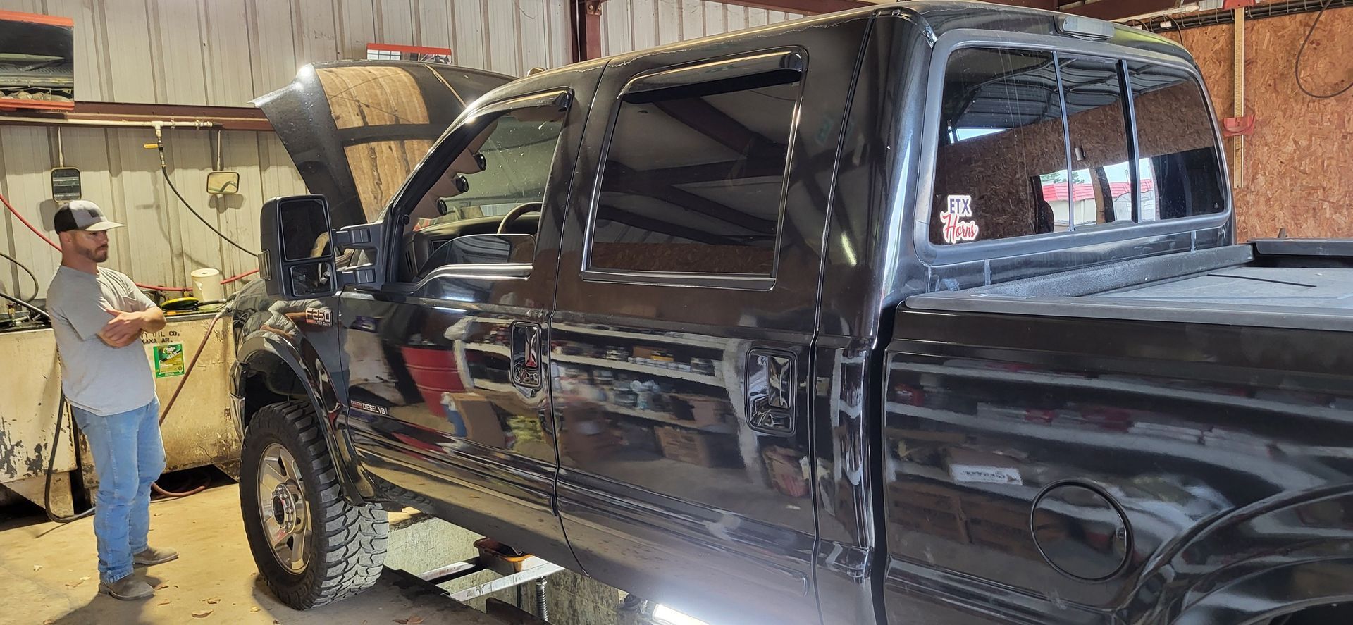 Black pickup truck in a garage with its hood open, while a man stands beside it and looks on.
