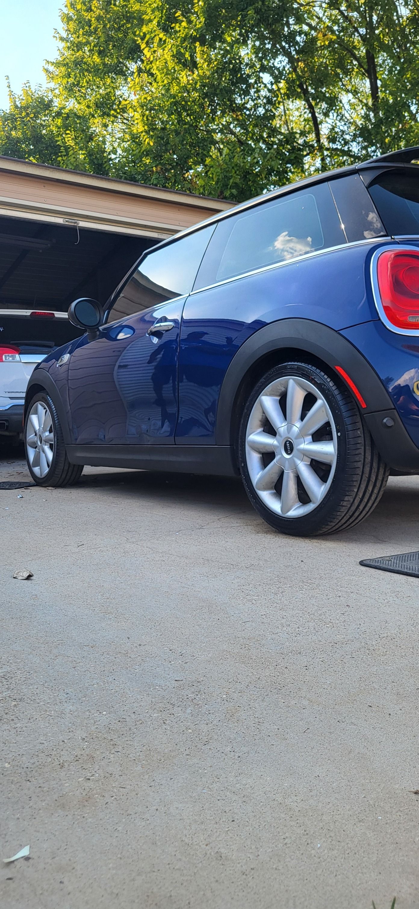 Blue hatchback car parked in a driveway beside a garage, viewed from the rear side.