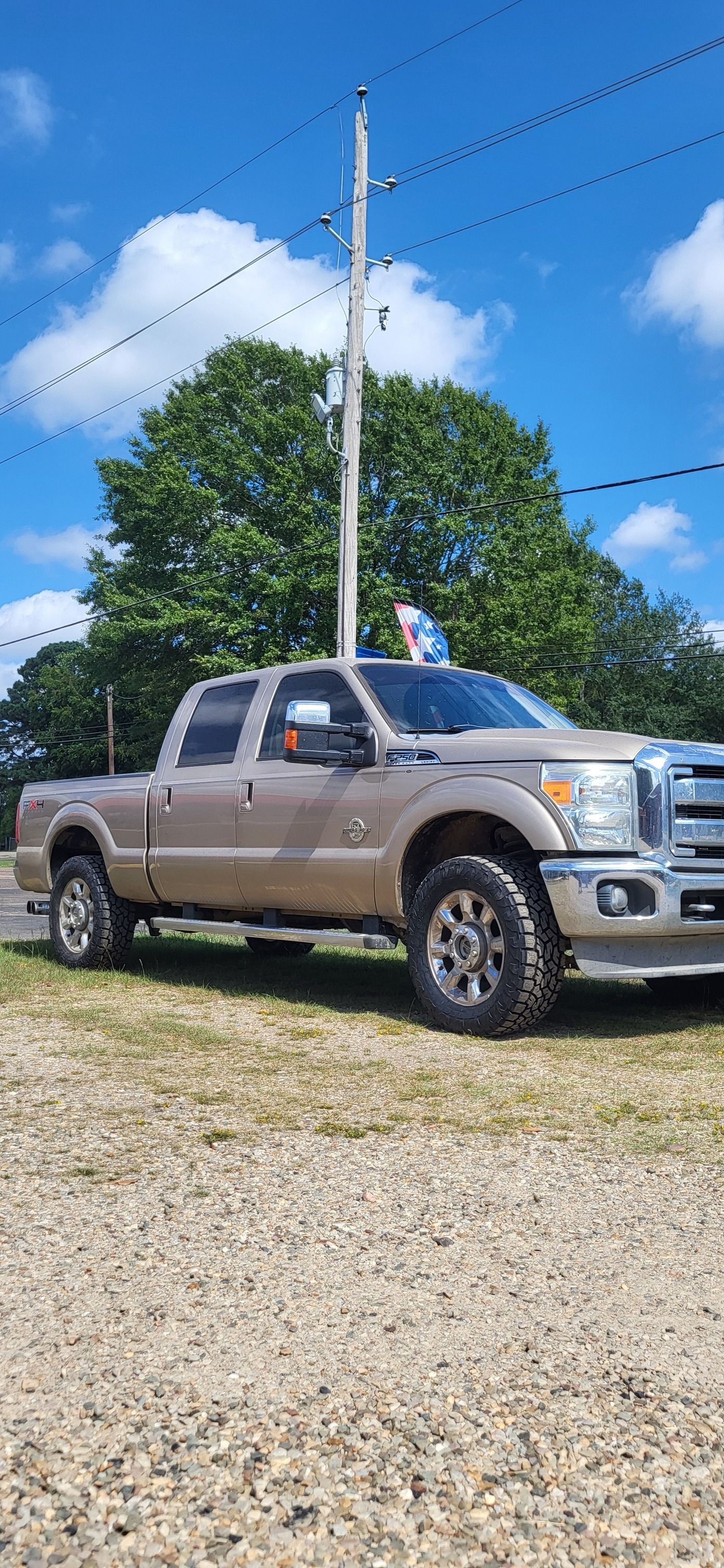 Tan pickup truck parked on gravel under a blue sky near power lines and trees