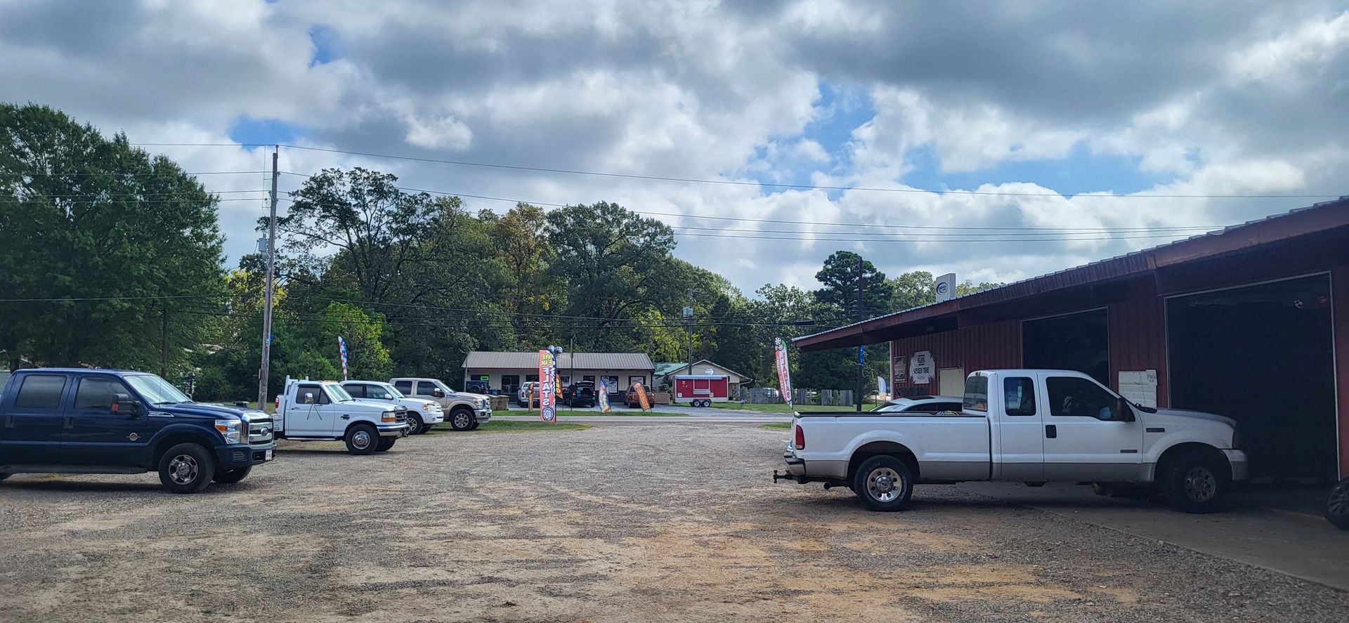 Rural parking lot with several vehicles, a long red-roofed building, and cloudy sky