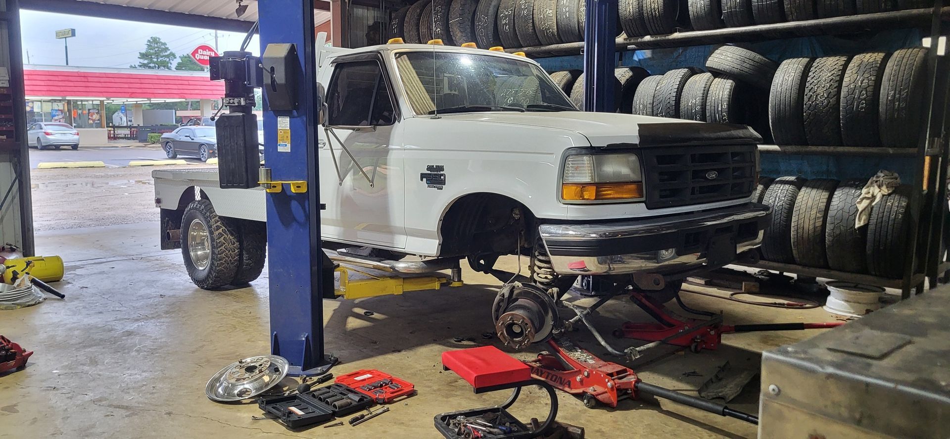 Auto shop interior with a white pickup truck on a lift and tires stacked along the wall, tools on the floor.