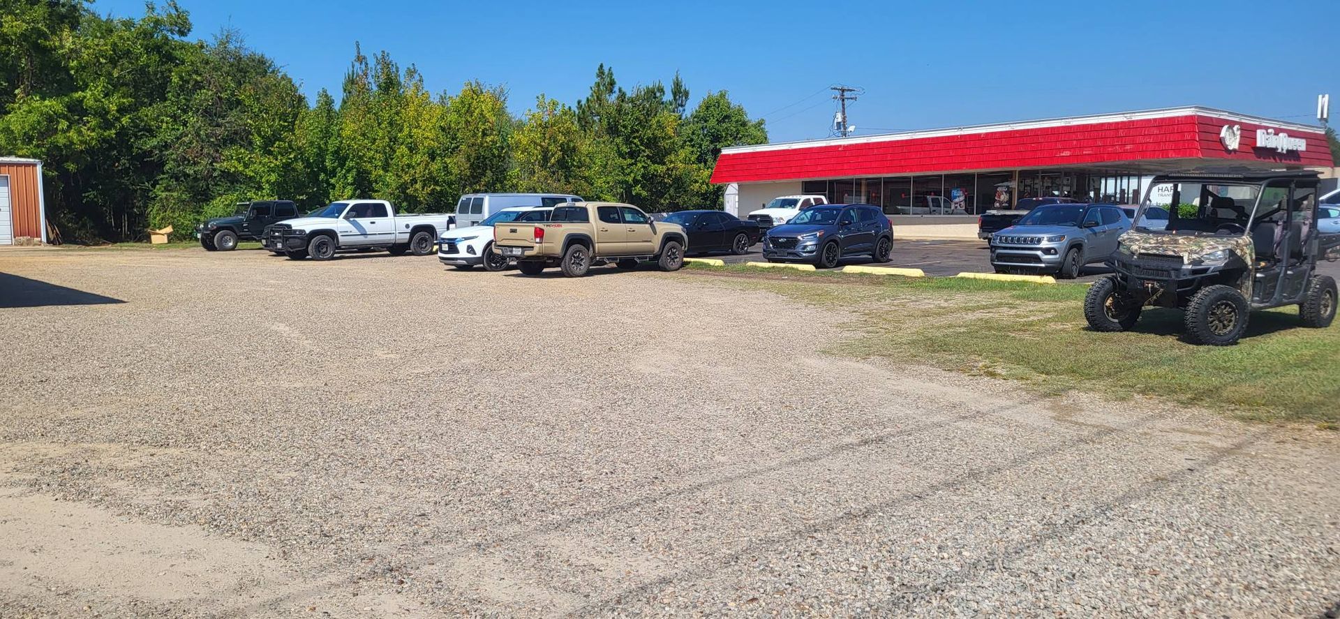 Gravel parking lot with several parked vehicles beside a red-roofed open shelter and trees under a clear sky