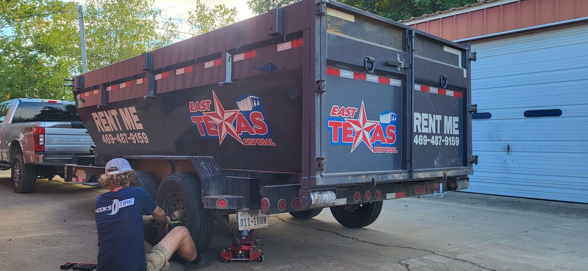 Tow truck operator working on a black trailer with red graphics outside a blue garage