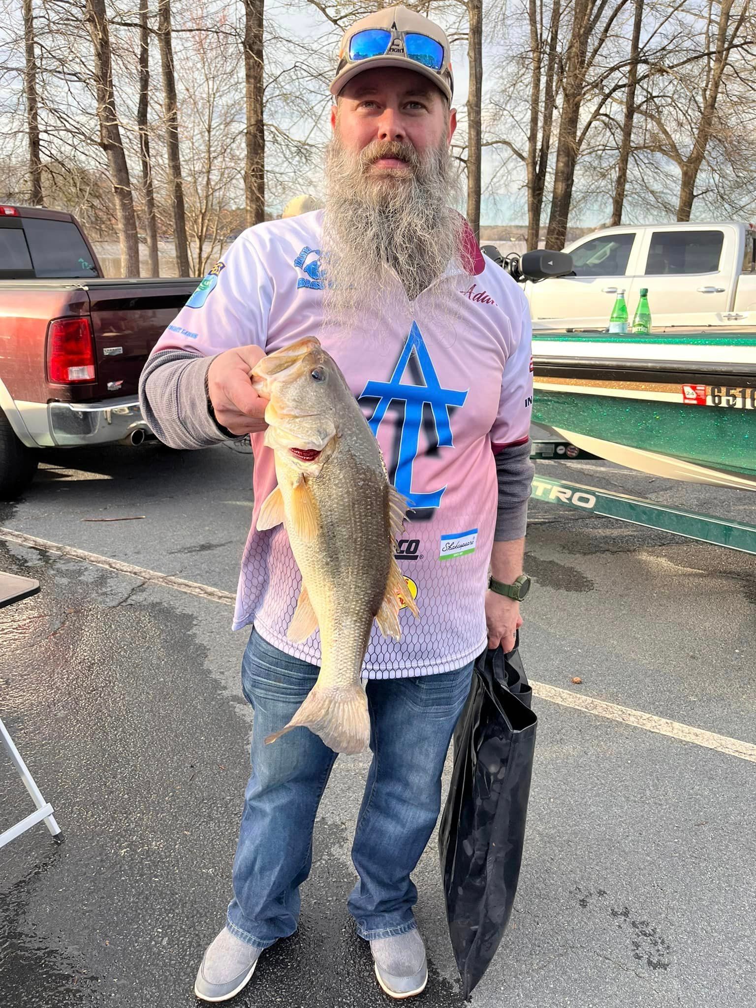 Man holding a large striped bass at a boat ramp. He's wearing a hat, long-sleeved shirt, and jeans. Trees in background.