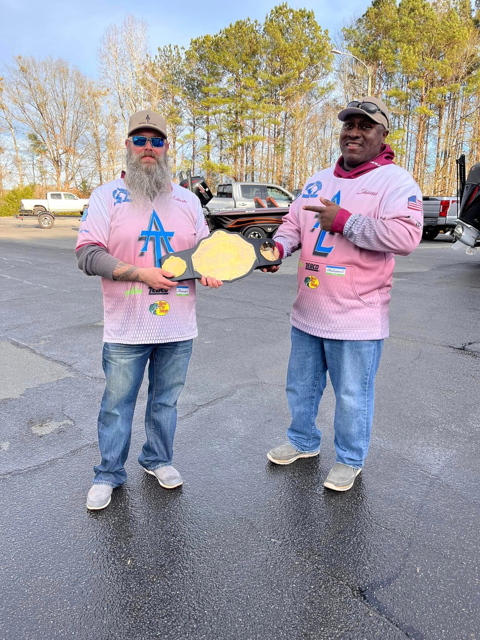 Two men in matching pink shirts holding a gold championship belt outdoors.