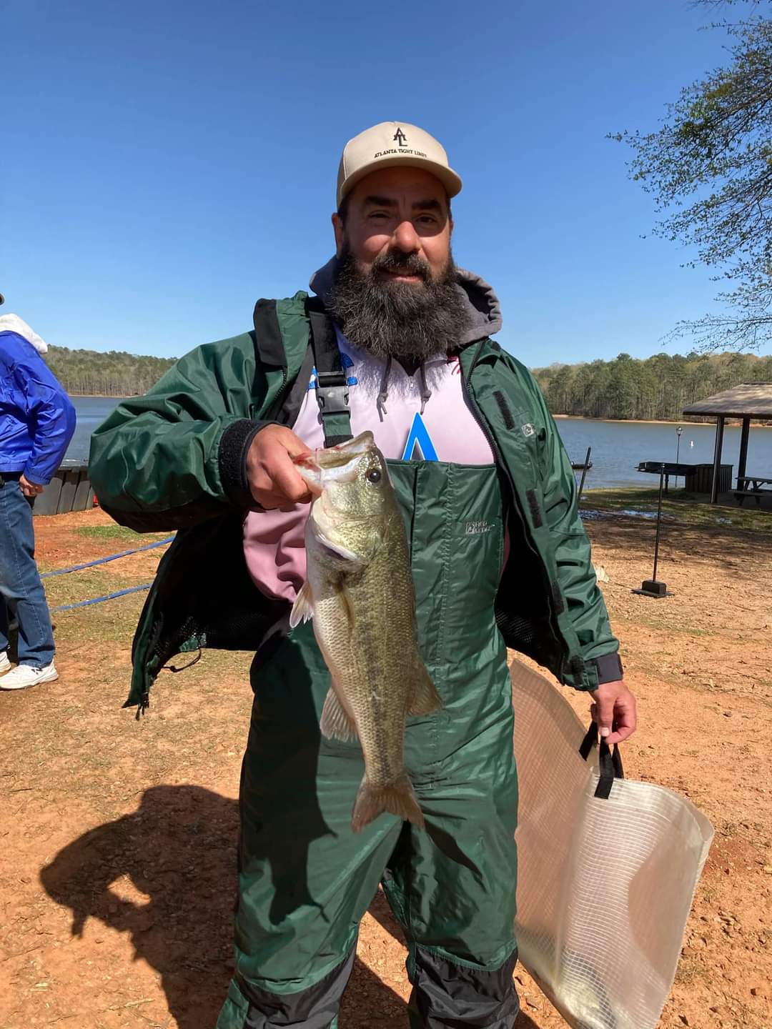 A man with a beard in waders holds a bass, with a lake and trees in the background.