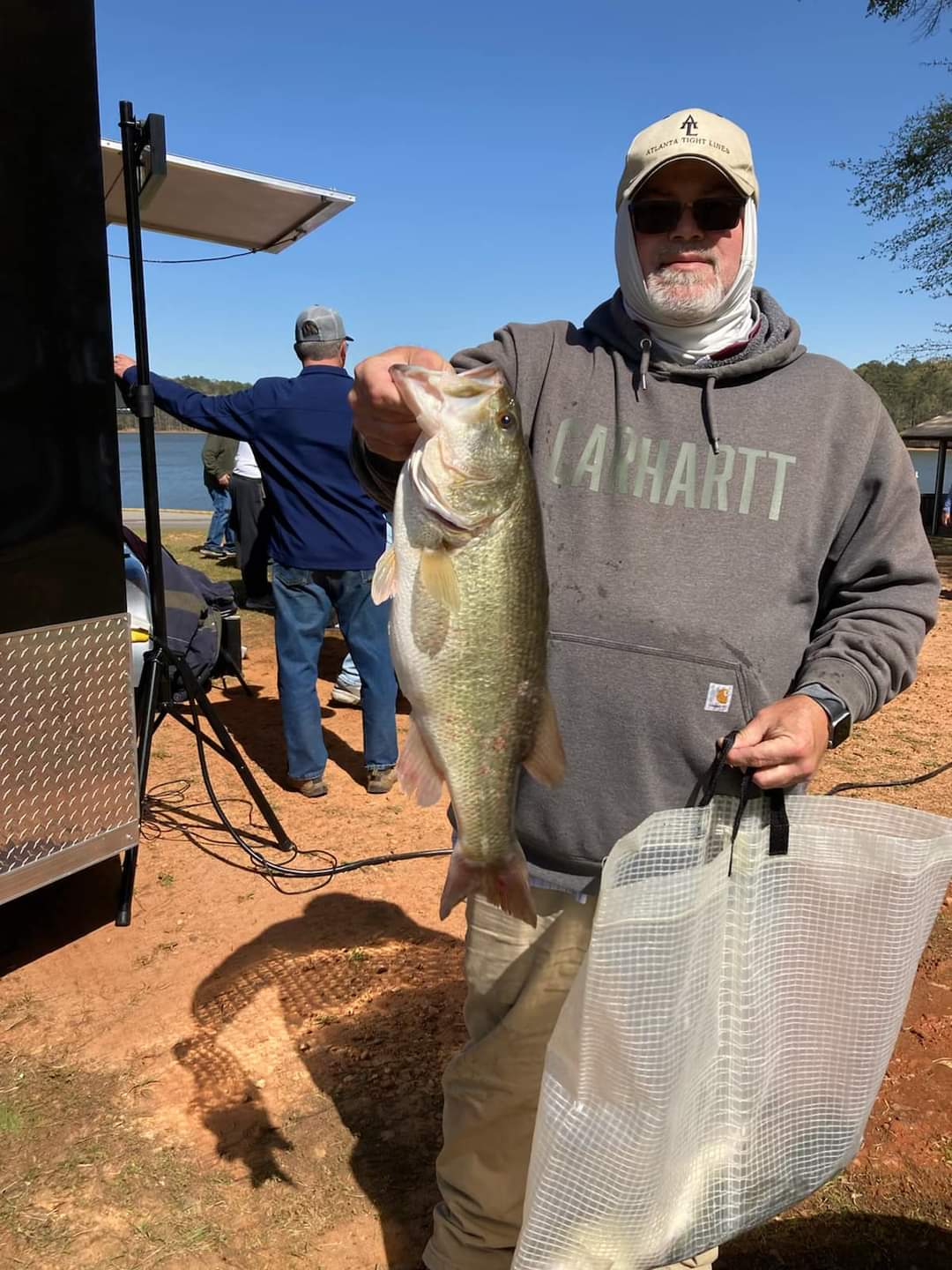 Man holding a large fish, wearing a Carhartt hoodie, near a lake on a sunny day.