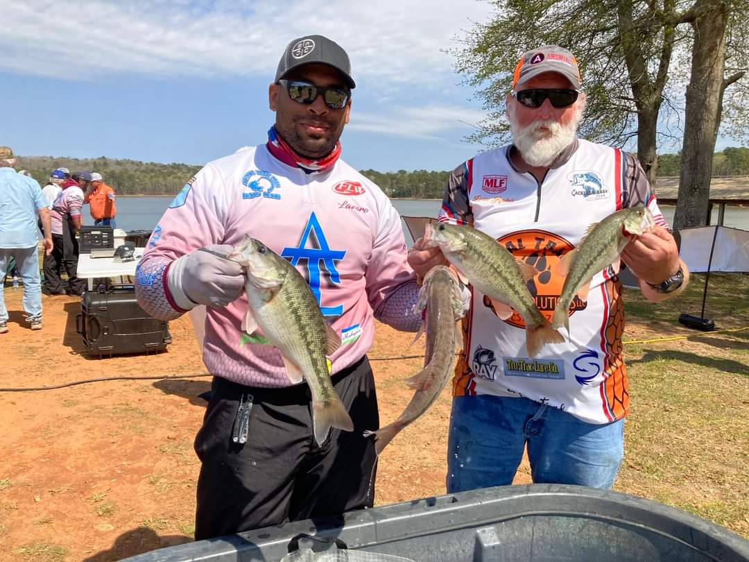 Two men holding fish at an outdoor fishing event. One has a blue symbol on his shirt, and the other has orange trim.