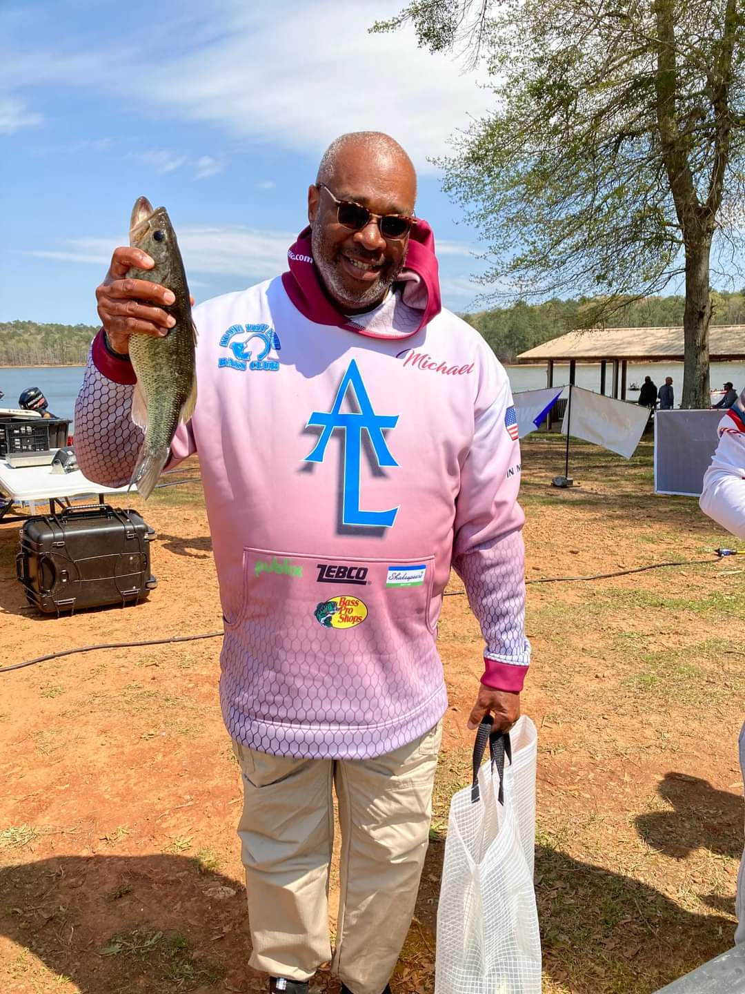 Man holding a fish, wearing a pink sweatshirt, standing outdoors near water.