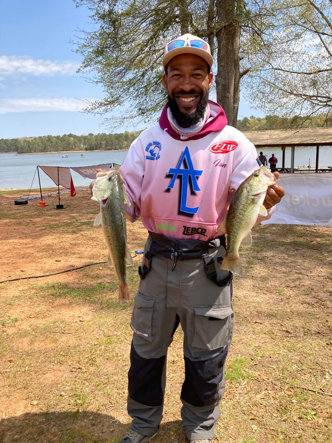 Man holding two bass; smiling, wearing fishing gear, lakeside setting.