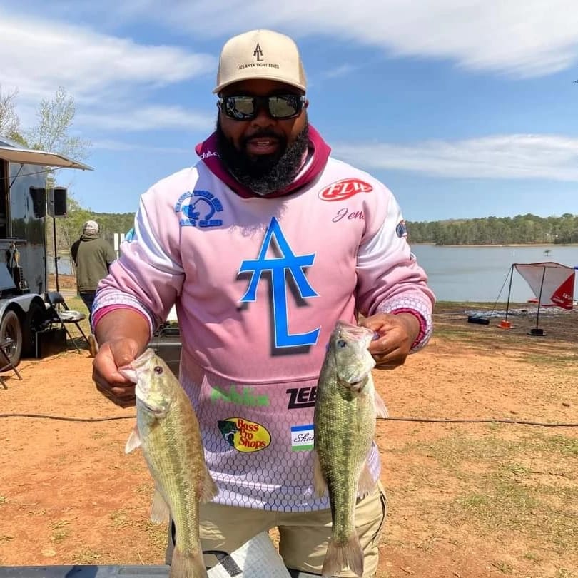 Man holding two bass fish, smiling, near a lake; wearing pink and white jersey, hat, and sunglasses.