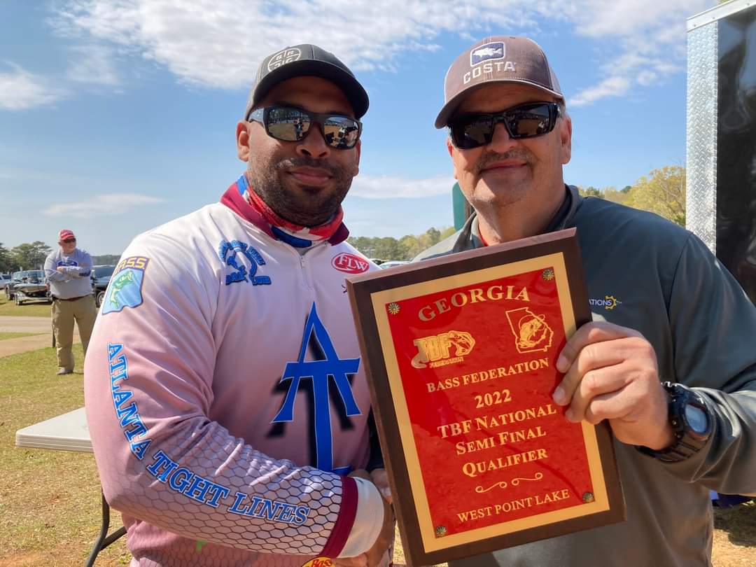 Two men holding a plaque, outdoors. One in a racing suit, other in a hat. Award says