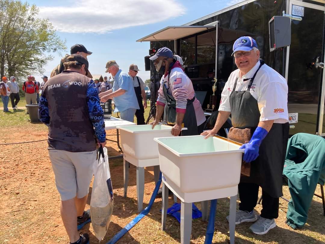 People washing fish at outdoor event; white sinks, mobile food trailer in background. Sunny day.